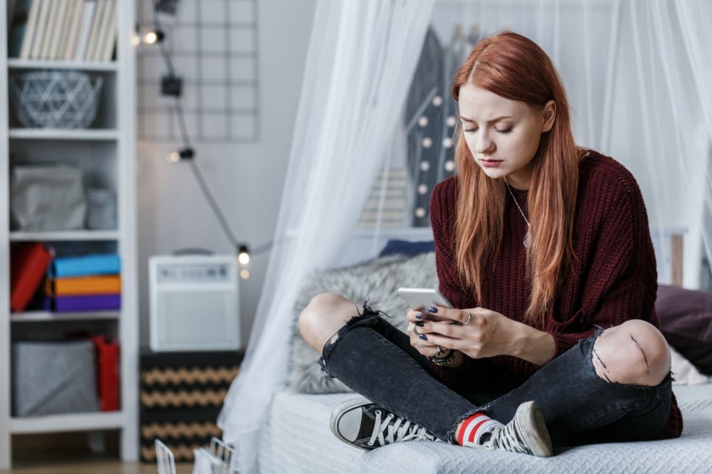 Upset teen sits on her bed while looking at her phone. She is looking at the nasty things others are saying about her friends. Therapyology offers group therapy for teen girls in west bloomfield, mi, teen girl therapy groups in west bloomfield, mi, and more.