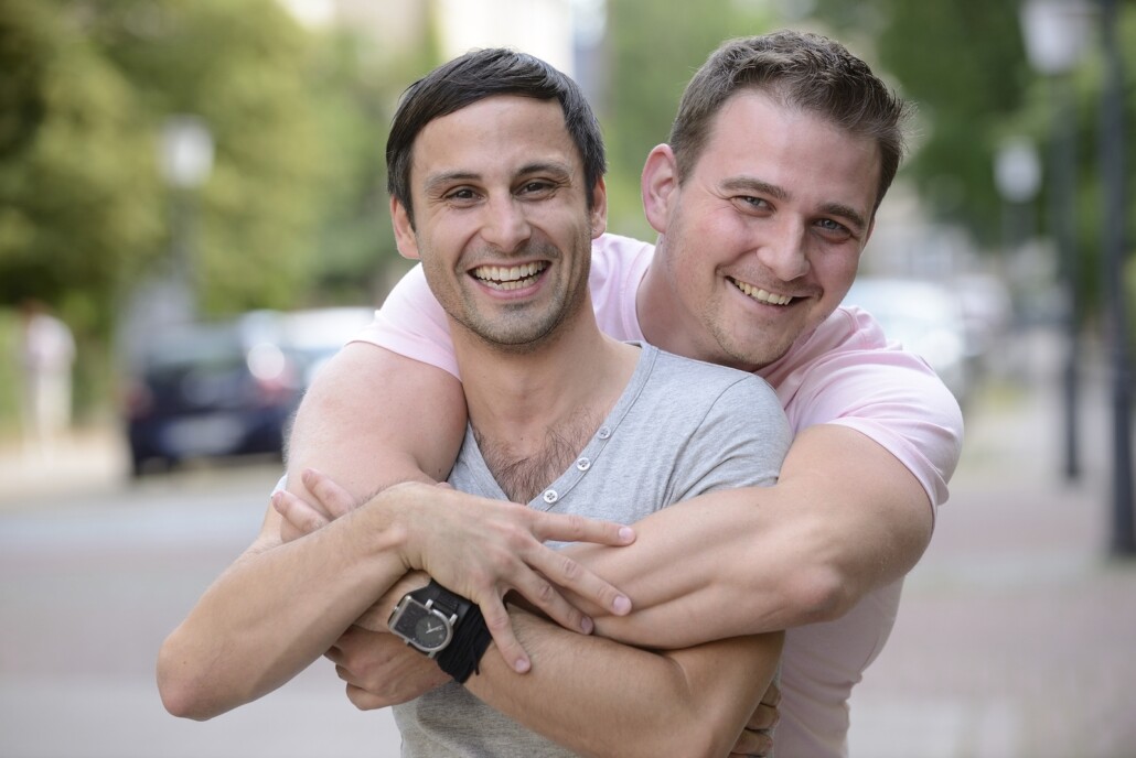 A gay couple stand outdoors as they look at the camera. A man is embracing his partner from behind, and both are smiling brightly. They are feeling good after meeting a lgbt ally in west bloomfield, mi at Therapyology. We offer lgbtq therapy in west bloomfield, mi, lgbt affirmative therapy in west bloomfield, mi, gender therapy in west bloomfield, mi, and more.