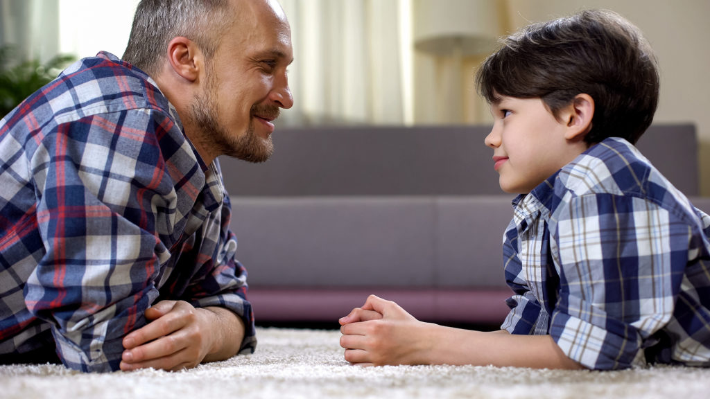 Man smiles at son as both lay on the carpet in front of one another. They are happy that they've found divorce therapy for children in West Bloomfield, MI. Therapyology offers therapy for teens of divorce, child divorce therapy, and more. Contact us for child therapy for divorce today.