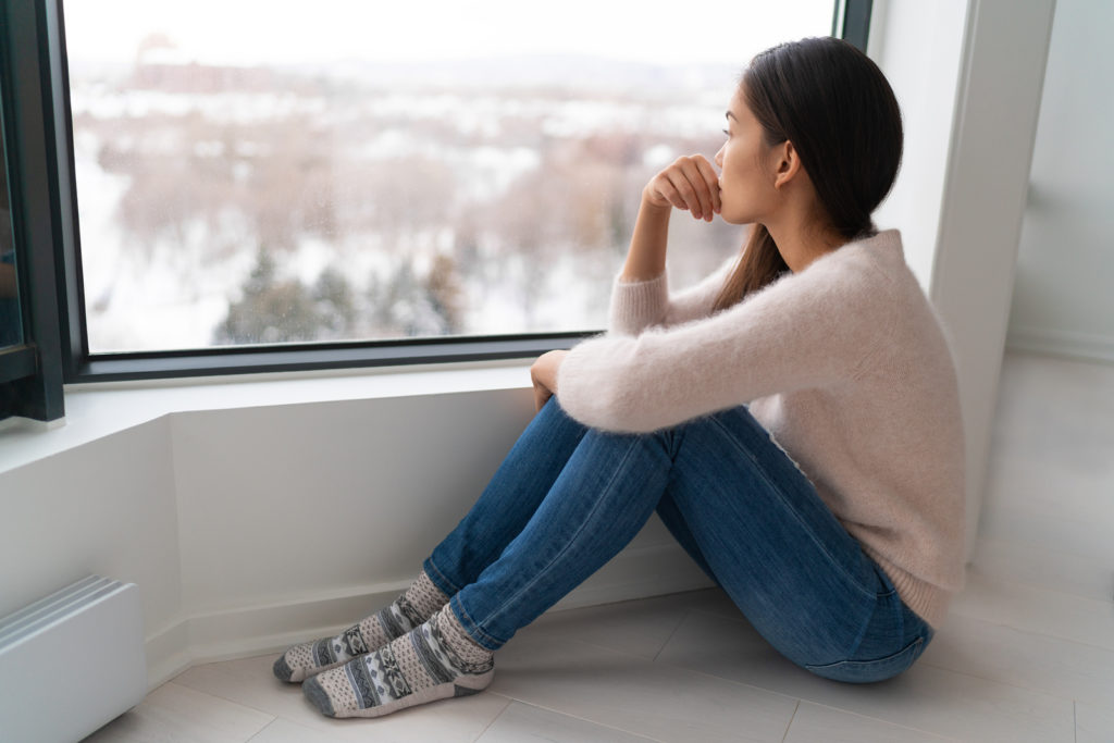 Young woman appears depressed as she sits on the floor looking out the window. She is thinking about past trauma. Therapyology offers trauma therapy in West Bloomfield, MI. Contact us for trauma counseling, trauma informed therapy, and more. Contact us to get in touch with a trauma therapist online in michigan.