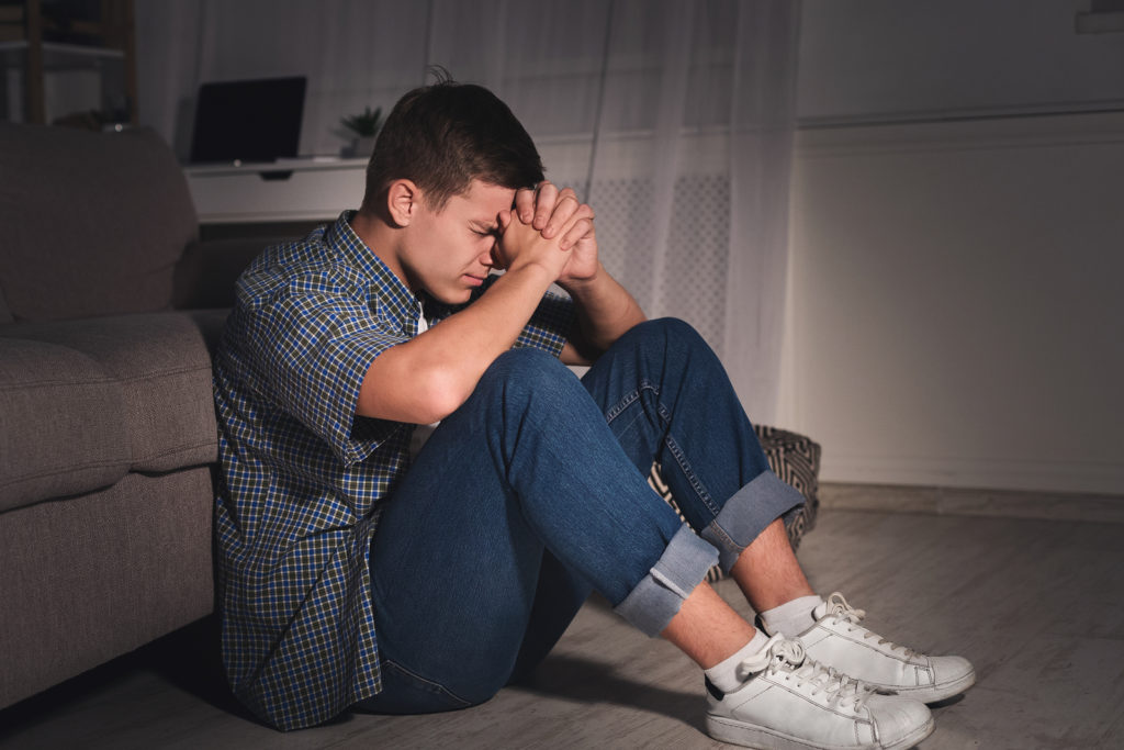 Teen sits on the ground with his head against his clenched hands. He is struggling with trauma, and has been working with a trauma therapist in West Bloomfield, MI. Therapyology offers online trauma therapy in Michigan, PTSD treatment, and more.