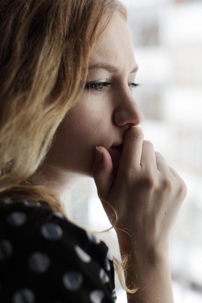 A close up of a woman holding her hand against her mouth while looking out a window. She appears worried about something as she is deep in thought. This could symbolize the fear and apprehension of a young adult. We offer transitions counseling in West Bloomfield, MI for young adults adapting to change. Contact a young adult therapist for young adult therapy and other services.