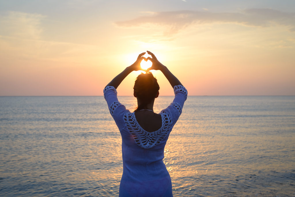 Woman at the beach holds her hands above her head in a heart shape. Shining between her hands, the sun can be seen setting over the water. This symbolizes the peace people feel after dealing with grief. We offer grief counseling in West Bloomfield, MI. If you're going through the stages of grief, contact a grief counselor for support!