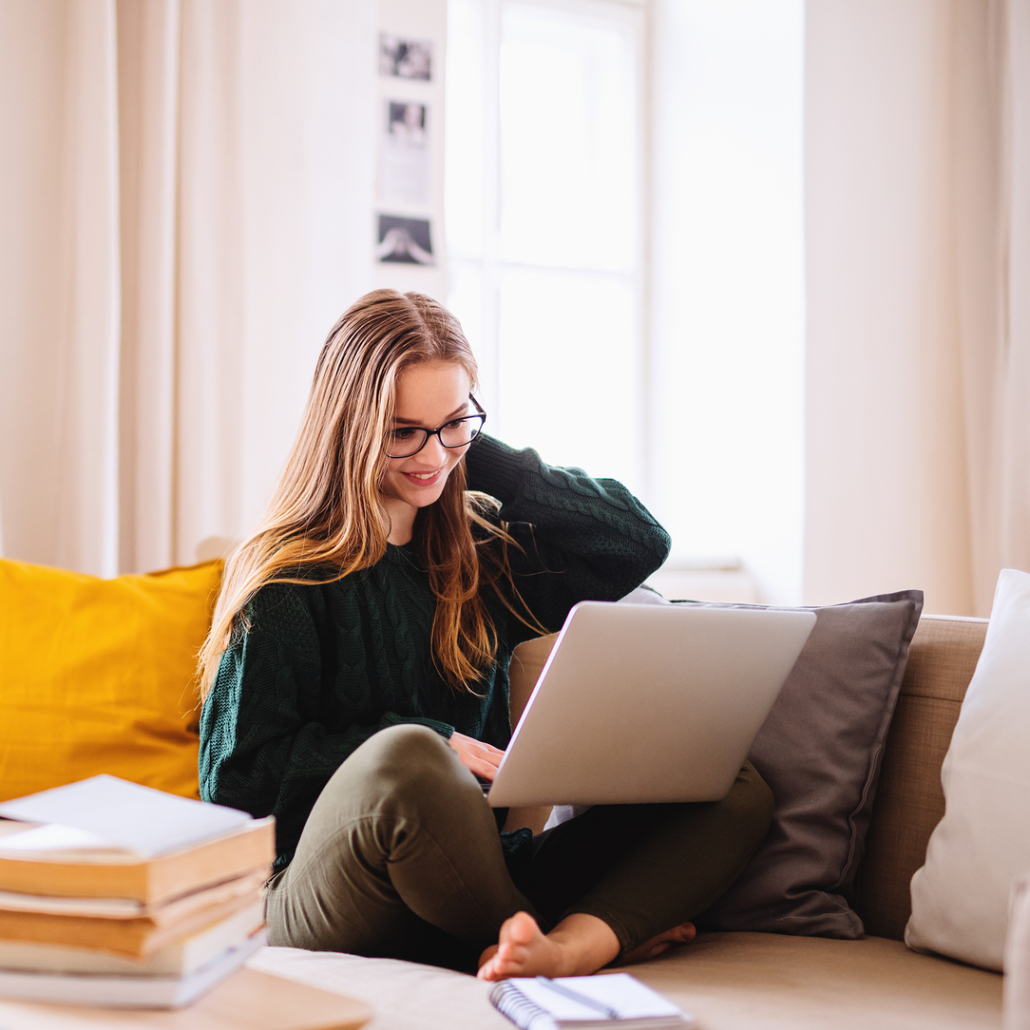 A young woman smiles as she sits on her sofa with a laptop in hand. You can meet with an online therapist in California from the comfort of home. Contact Therapyology to learn more about online counseling for teens and other services.