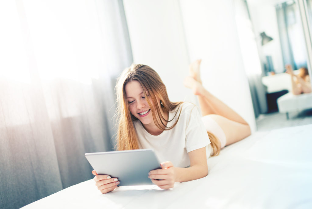 A teen smiles at her tablet as she lays on her stomach. This could represent the comfort of online therapy in California. An online therapist can share more info about online trauma therapy, online therapy for teens, and other services.