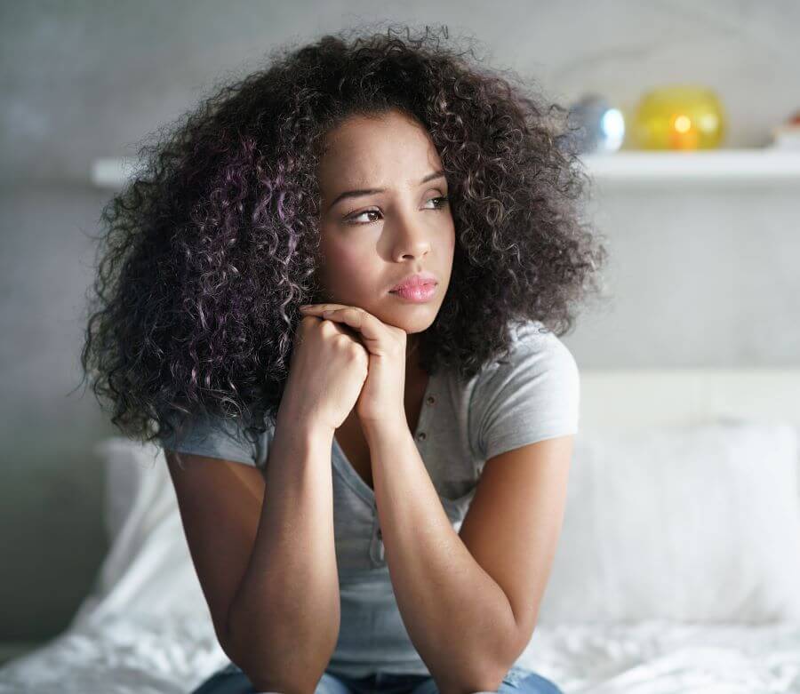 A sad young woman sits alone in bedroom. This symbolizes the isolation that can come from the stages of grief. Therapyology offers grief counseling in West Bloomfield, MI. Contact a grief counselor for support dealing with grief.